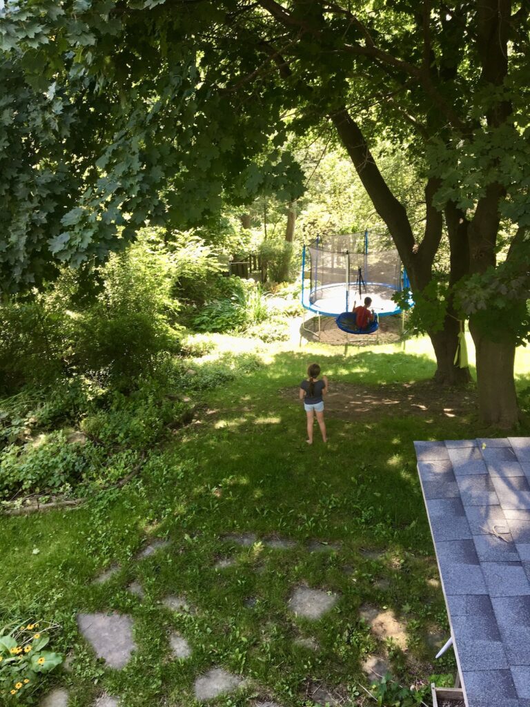 A child stands in a green backyard under tall trees, looking toward a trampoline where another child plays. A child stands in a green backyard under tall trees, looking toward a trampoline where another child plays.